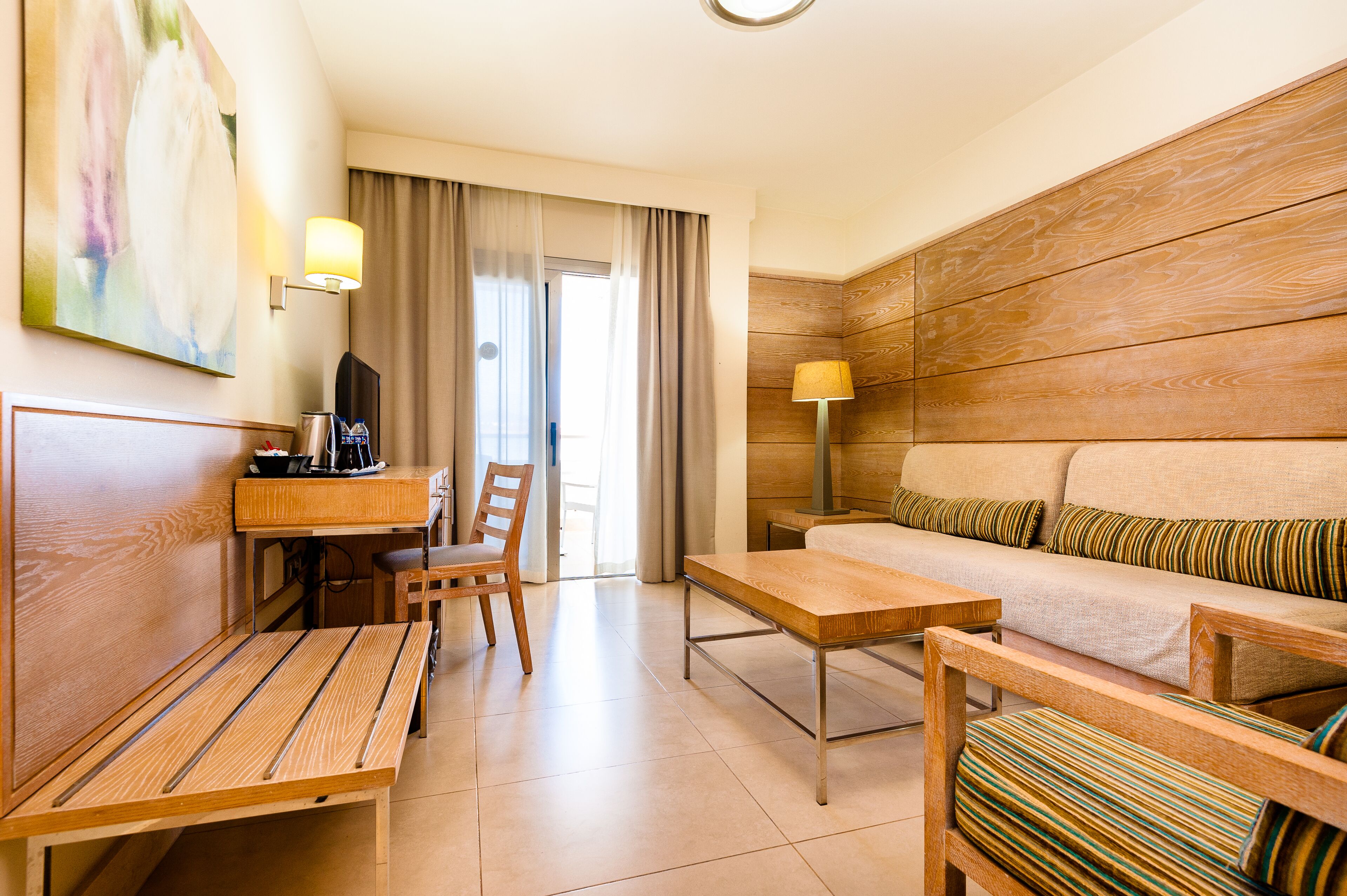 A hotel room with a wooden accent wall, a sofa with striped cushions, a dining table, a chair, a desk with a lamp, and a television.