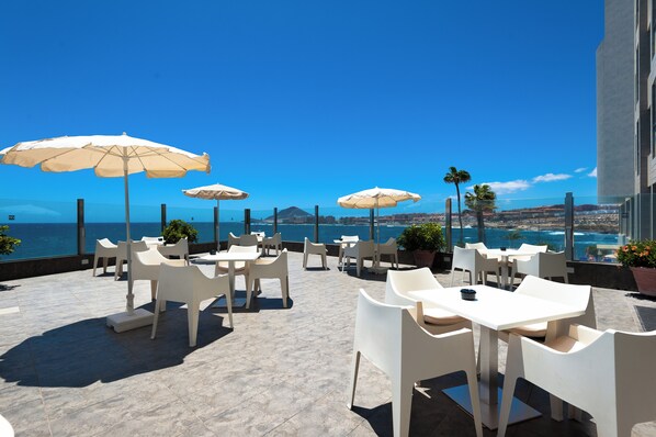 A terrace with white chairs and tables, white umbrellas, and a view of the ocean.