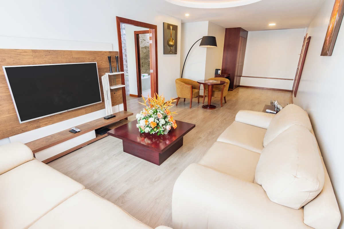A modern living room with a flat-screen TV, a wooden coffee table with a floral centerpiece, a sofa, and a dining area with a table and chairs.