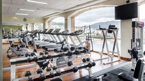 A gym with treadmills, dumbbells, and weight machines, large windows with a city view, and a mounted TV. - Sonesta Hotel Loja (Loja)