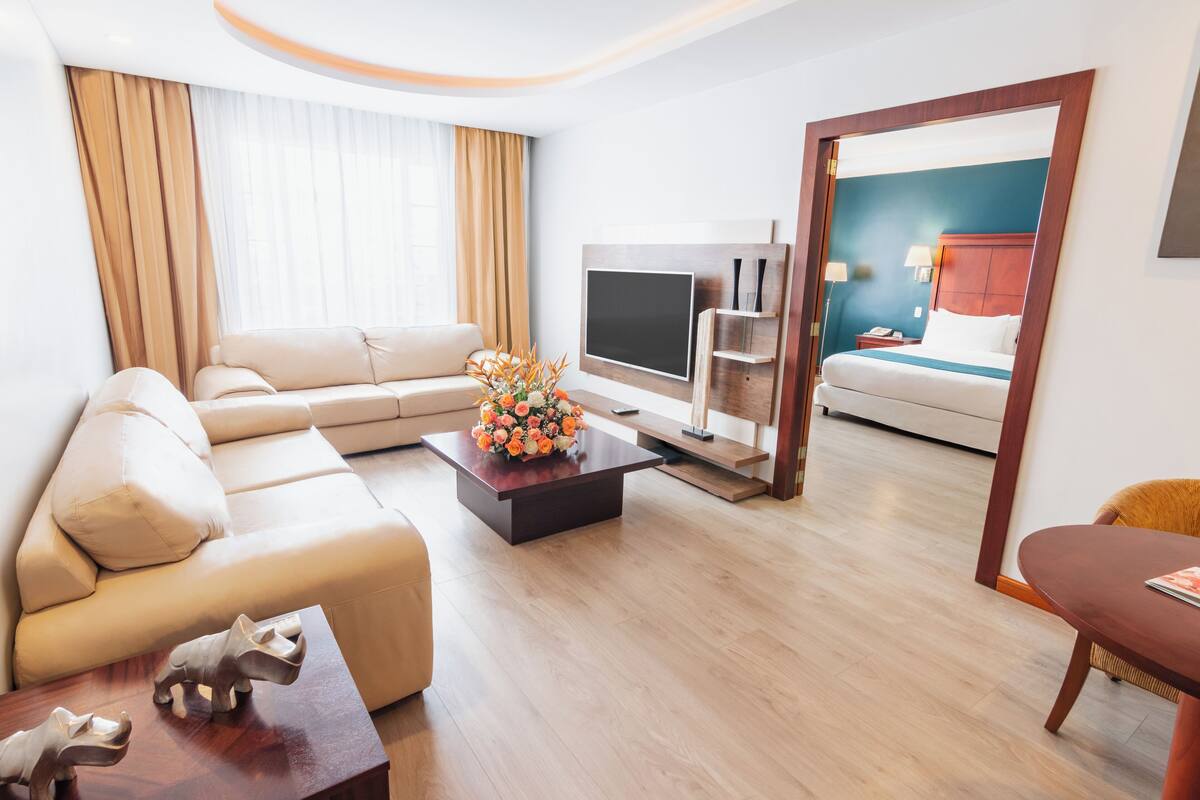 Ein modernes Hotelzimmer mit einer Couch, einem Couchtisch mit floraler Dekoration, einem Flachbildfernseher und einem Bett mit Holzrahmen.