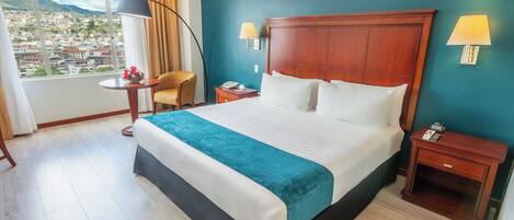 Habitación de hotel con una cama grande, dos mesitas de noche, una butaca, una silla, una pequeña mesa con lámpara y vistas a un paisaje urbano a través de un ventanal.