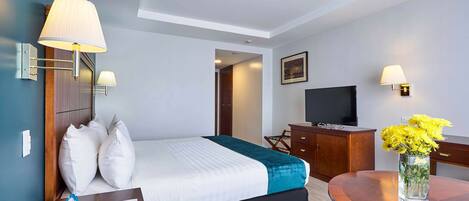 A hotel room with a bed, bedside table, lamp, desk, chair, and a TV.