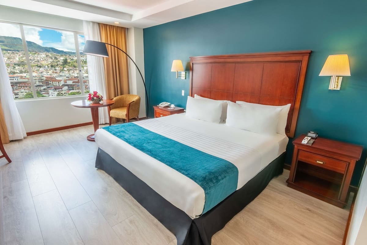 Ein Hotelzimmer mit einem großen Bett, Blick auf die Stadt, einem runden Tisch mit Stuhl und zwei Nachttischlampen.