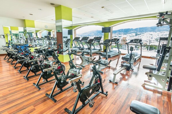 A gym with multiple exercise machines, a wooden floor, and a view of a cityscape through large windows. - Sonesta Hotel Loja (Loja)