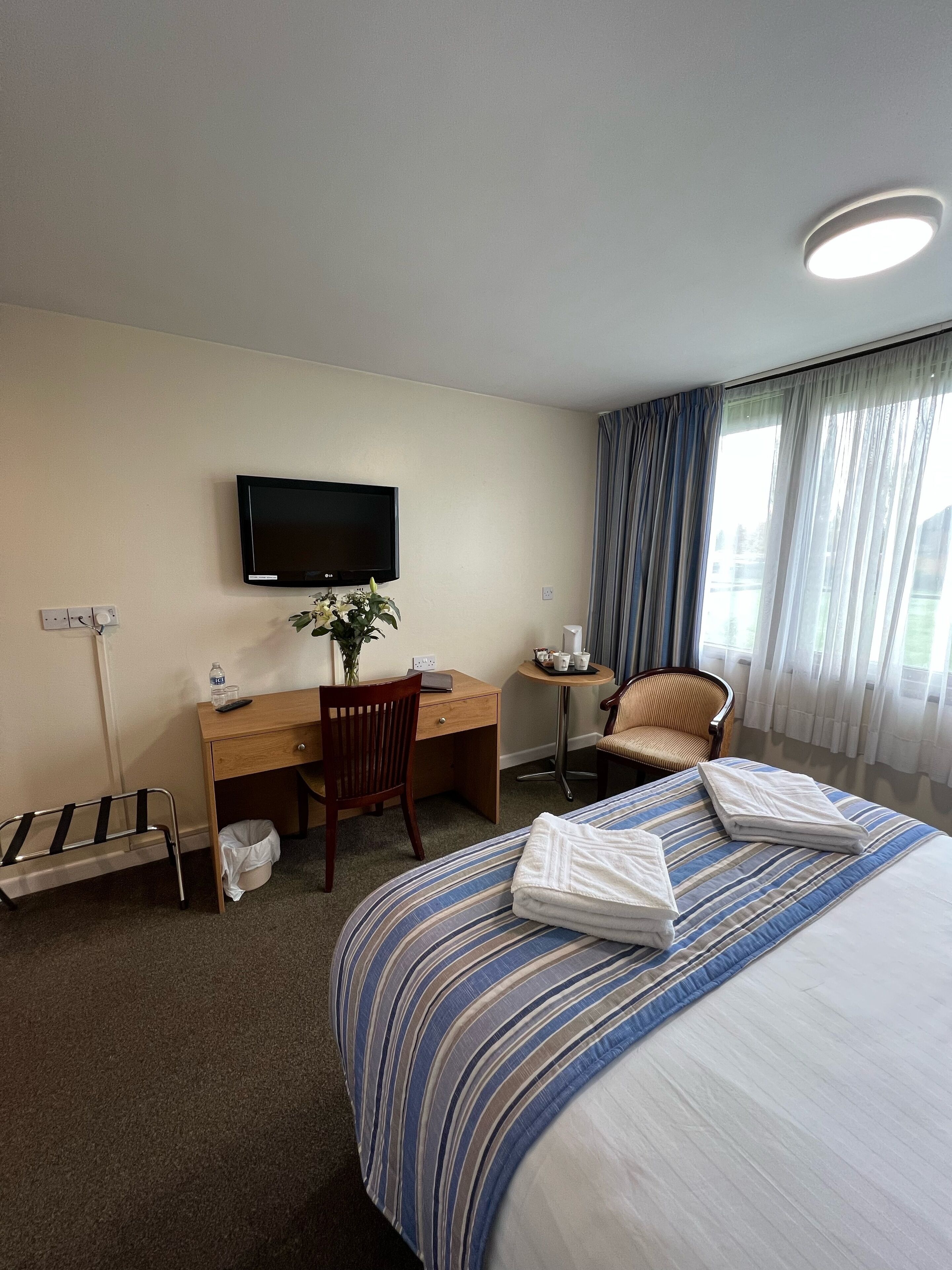 Habitación de hotel con cama, escritorio, silla, televisor y ventana con cortinas.