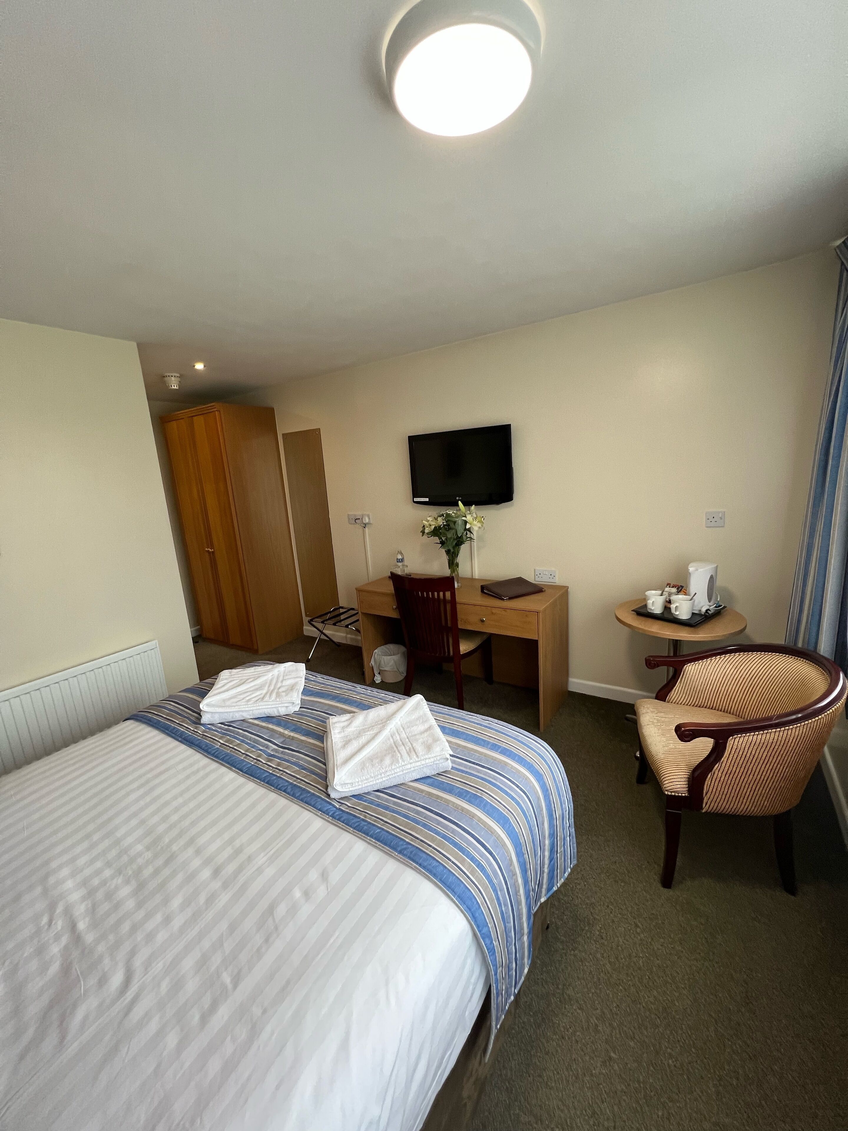 A hotel room with a bed, a desk, a chair, a TV, and a small table with a pitcher and cups.