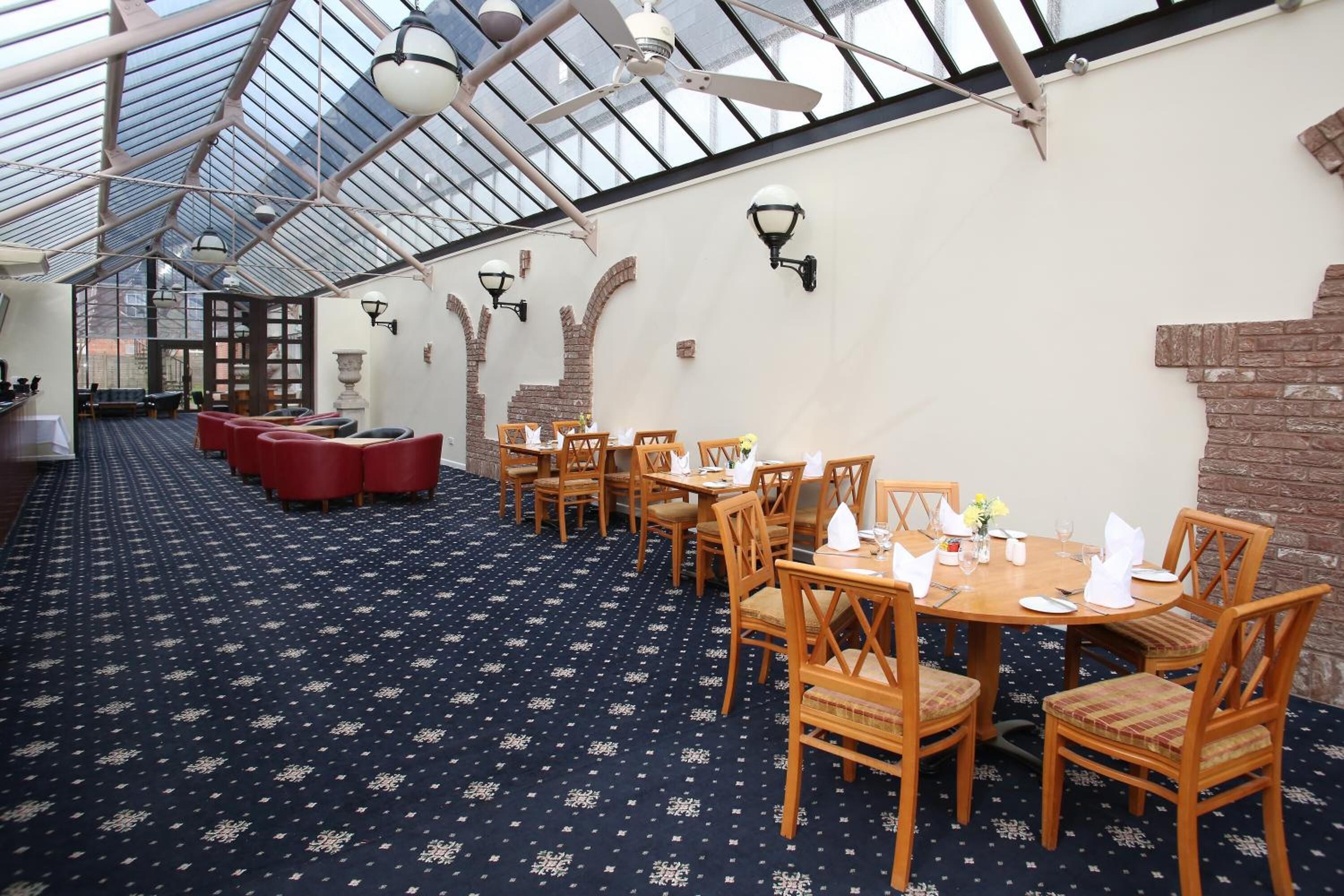 Ein Restaurant mit hohen Decken und Glasdach, Holztischen und -stühlen sowie einem gemusterten Teppich.