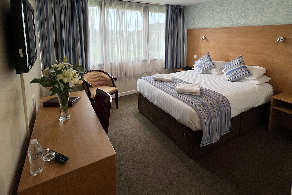 Habitación de hotel con cama, escritorio, silla, televisor y ventana con cortinas.