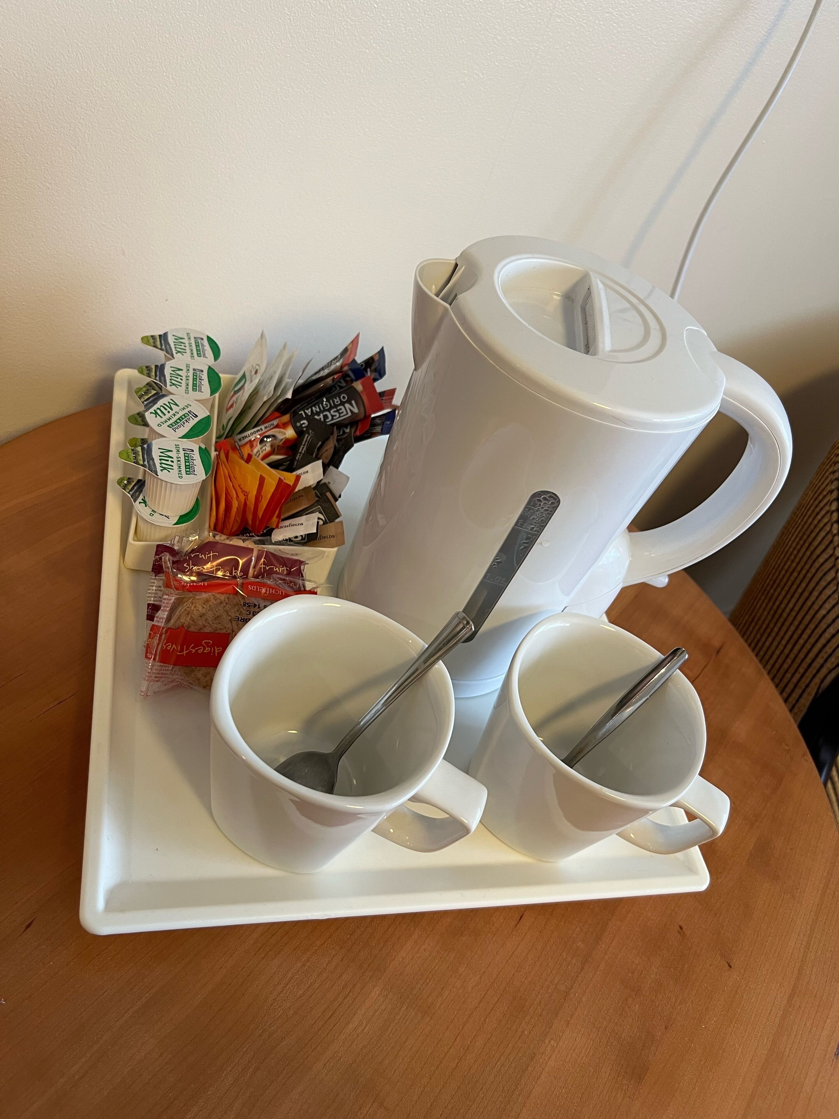 A tray with two white cups, a larger mug, and a spoon. There are also some packaged snacks and a small container.