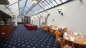 Un restaurante con techo alto y tragaluces, mesas y sillas de madera, y una alfombra estampada.