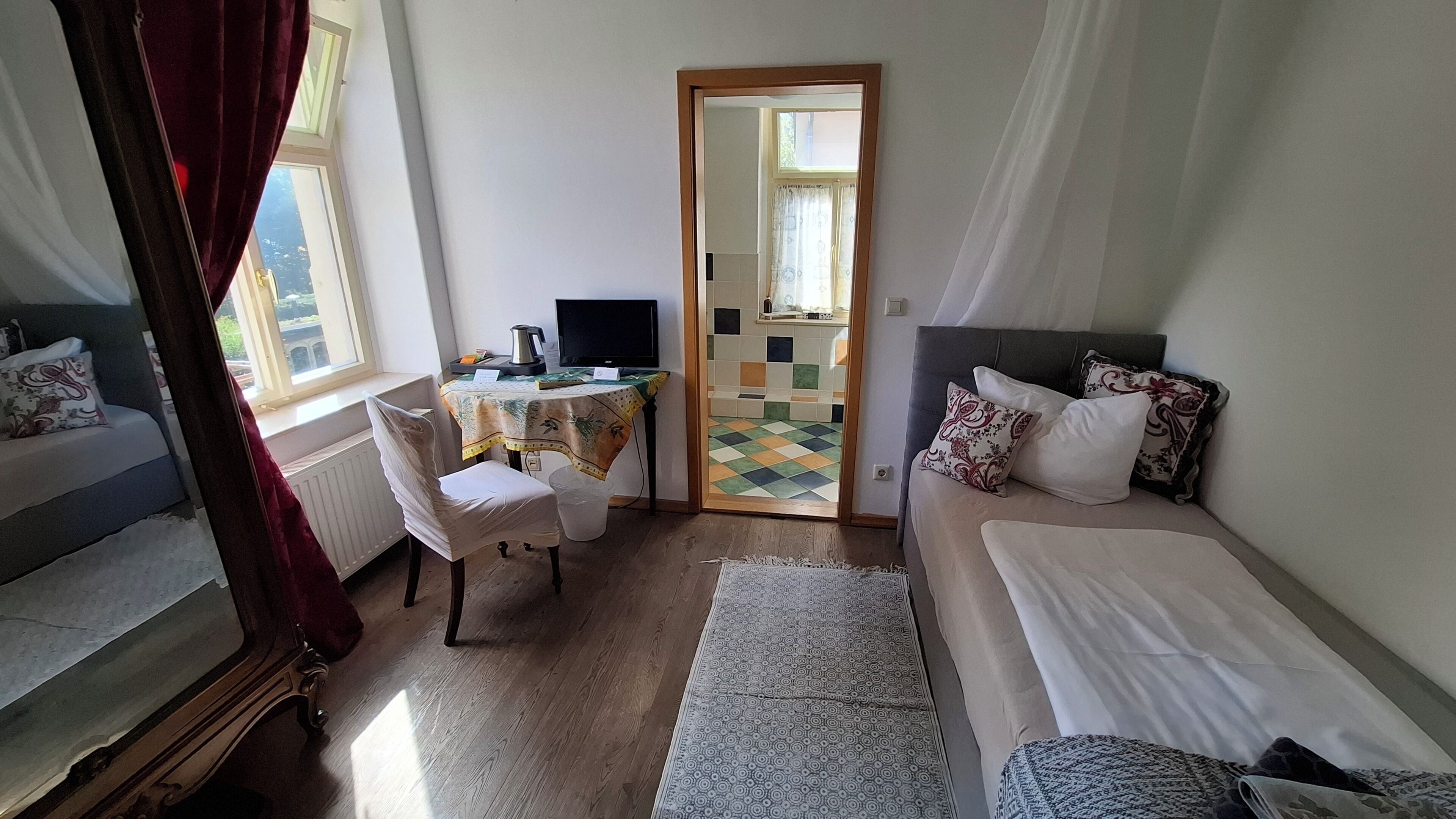 Una habitación con cama, escritorio, silla y vista a un área con baldosas a través de una puerta abierta.