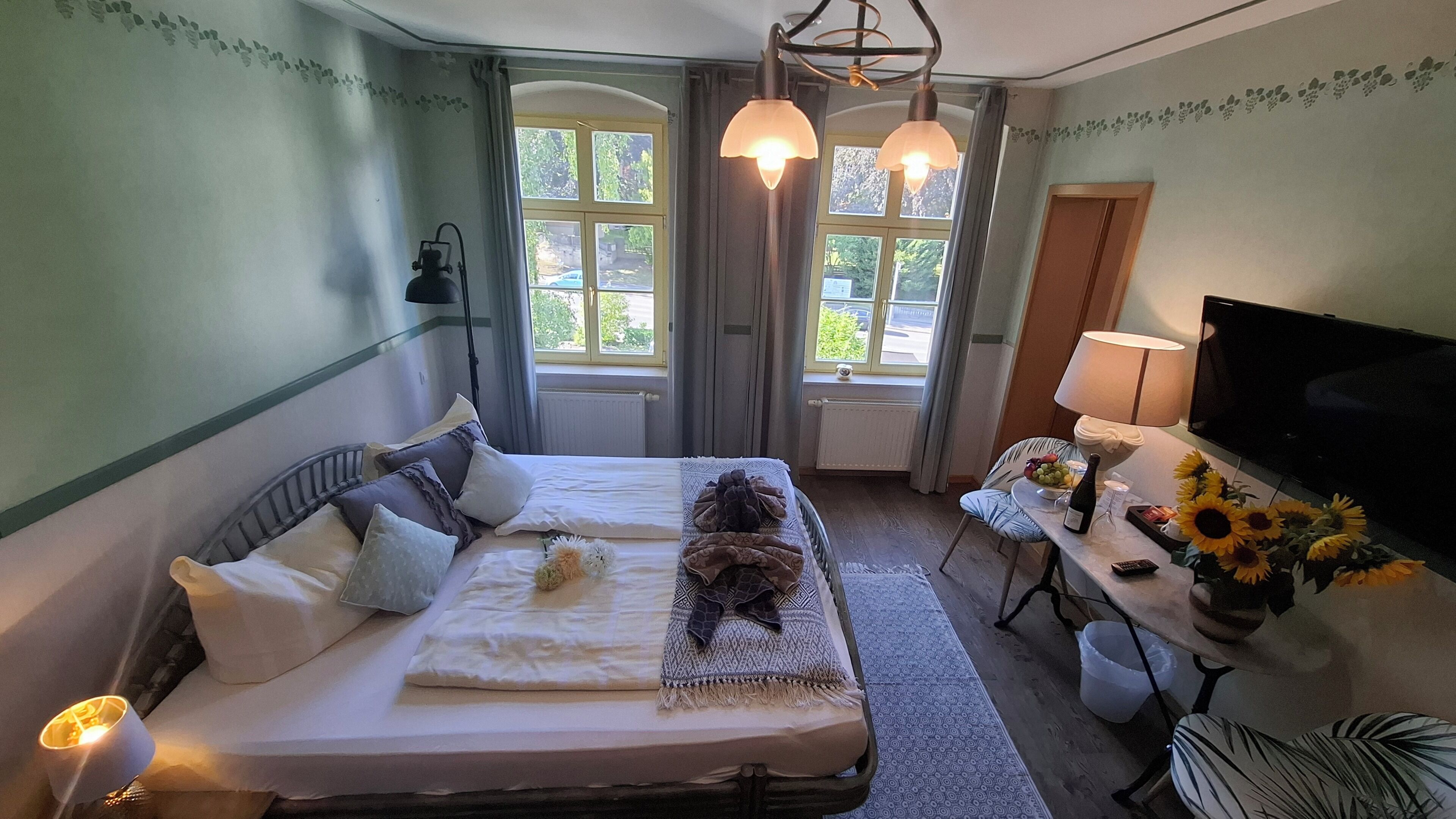 Ein Hotelzimmer mit einem Bett, zwei Fenstern, einem Fernseher, einem Tisch mit einer Vase voller Sonnenblumen und einer Lampe.