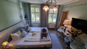 Una habitación de hotel con cama, dos ventanas, televisor, una mesa con un florero de girasoles y una lámpara.