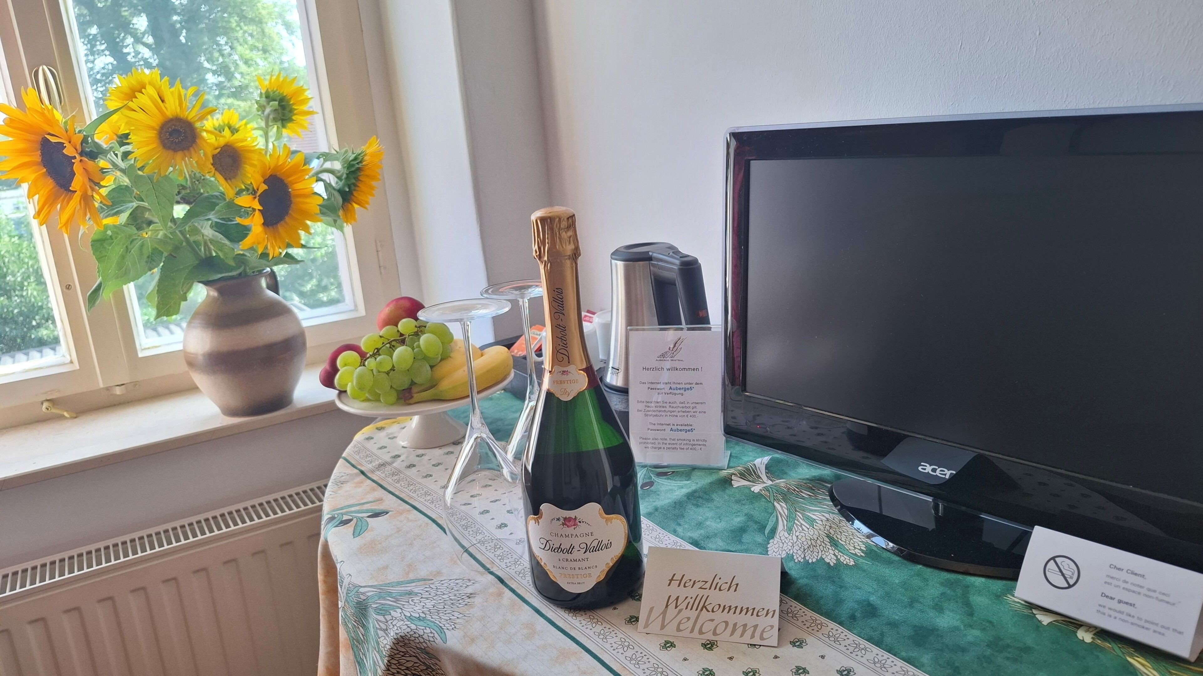 A table with a bottle of champagne, a vase of sunflowers, a fruit bowl, and a sign welcoming guests.