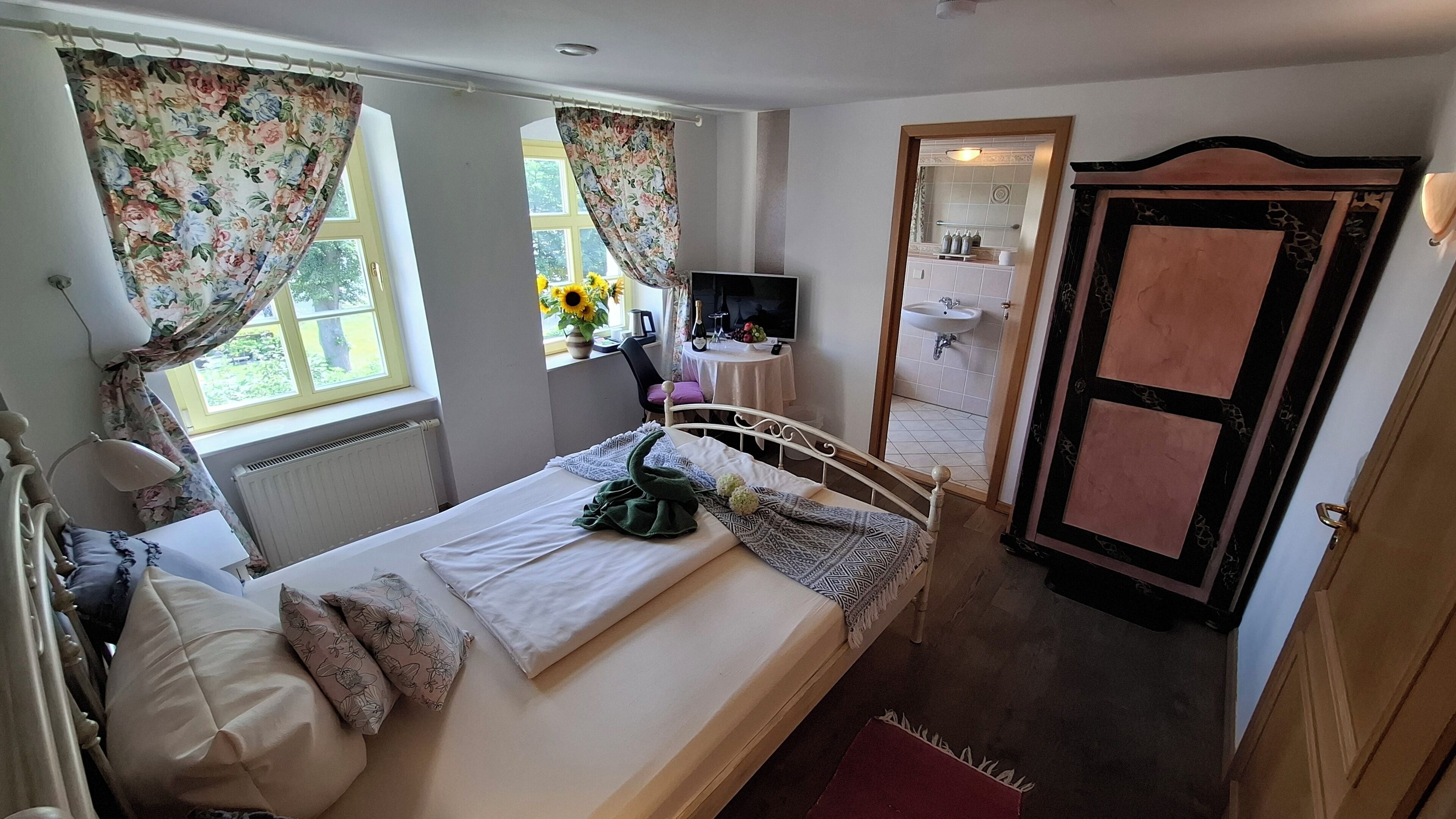 Una camera da letto con un letto, tende, un comodino, una sedia, una TV e un bagno visibile attraverso una porta aperta.