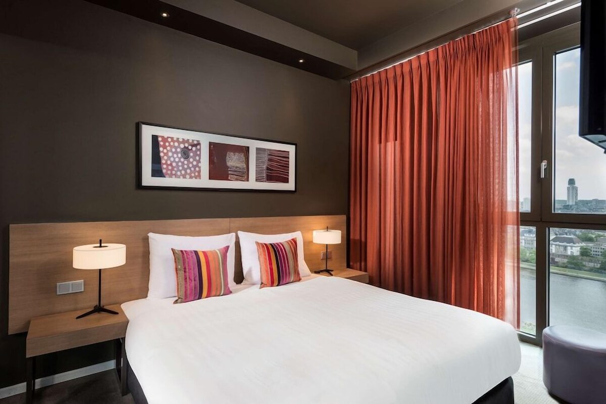 A hotel room with a large bed, two bedside lamps, a headboard, and a window with curtains.