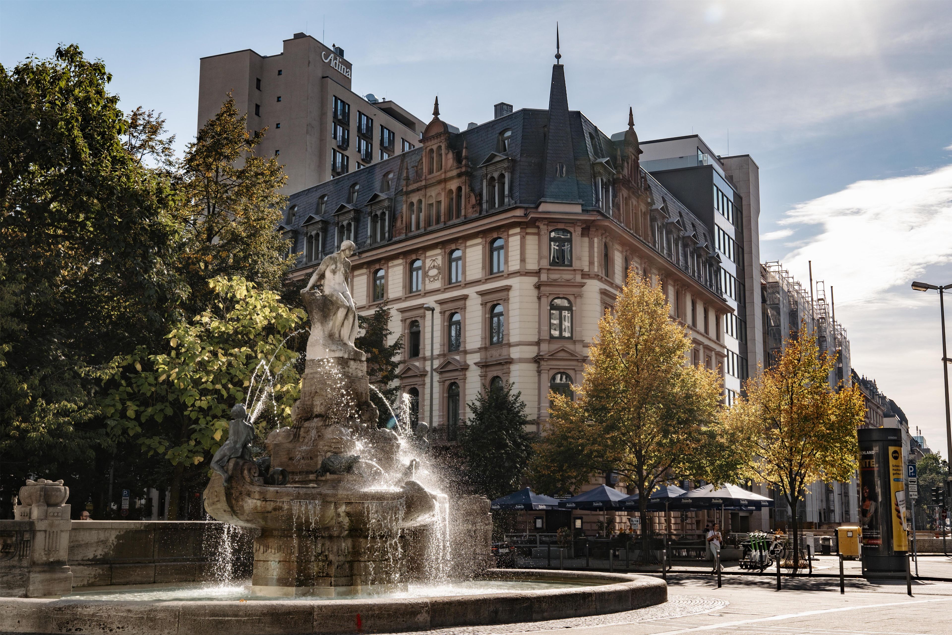 Ein Brunnen mit einer Statue in der Mitte, umgeben von Bäumen und Gebäuden.
