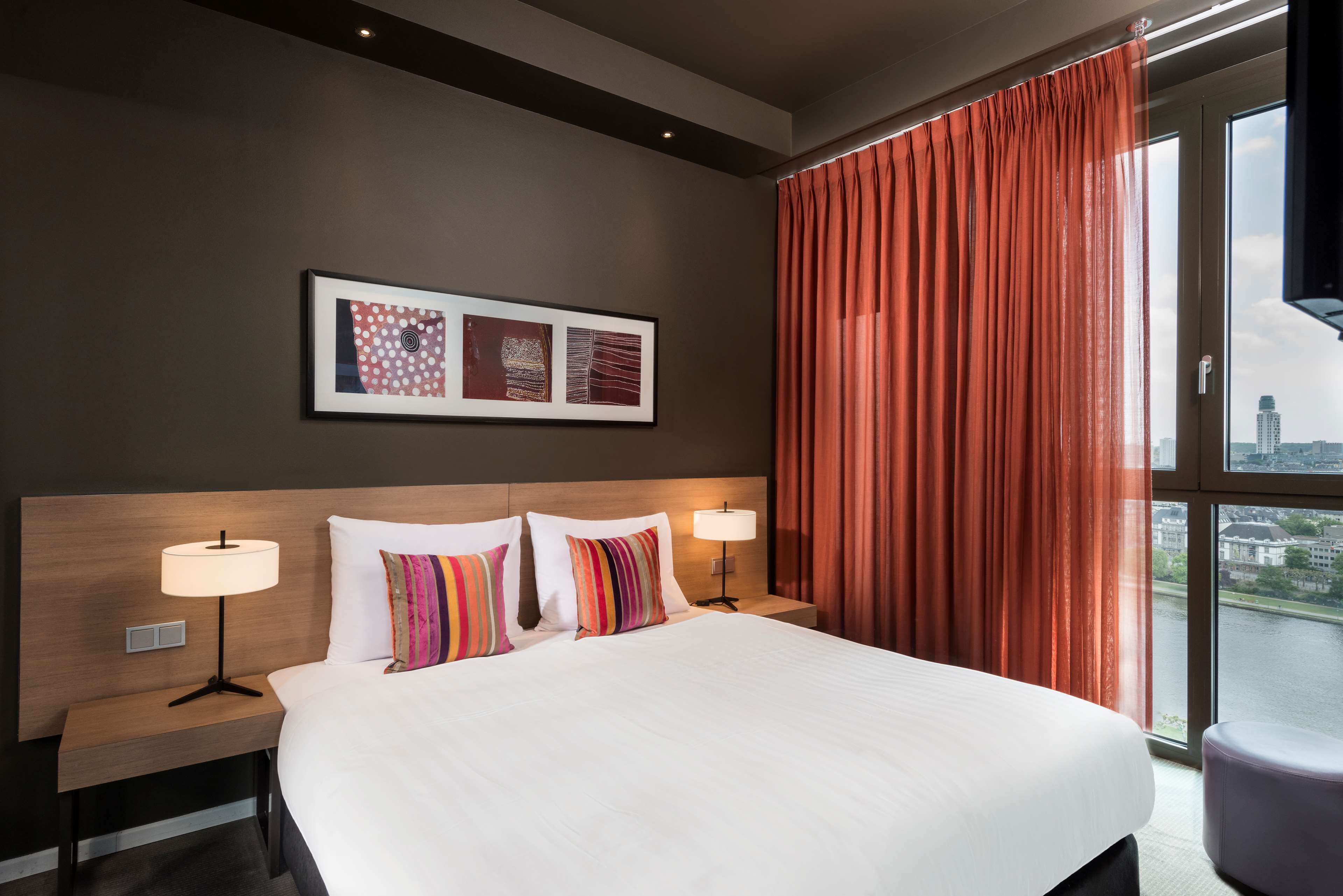 A hotel room with a large bed, two bedside tables with lamps, a view of a cityscape through a window, and artwork on the wall.