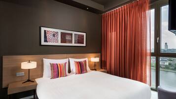 A hotel room with a large bed, two bedside tables with lamps, a view of a cityscape through a window, and artwork on the wall.