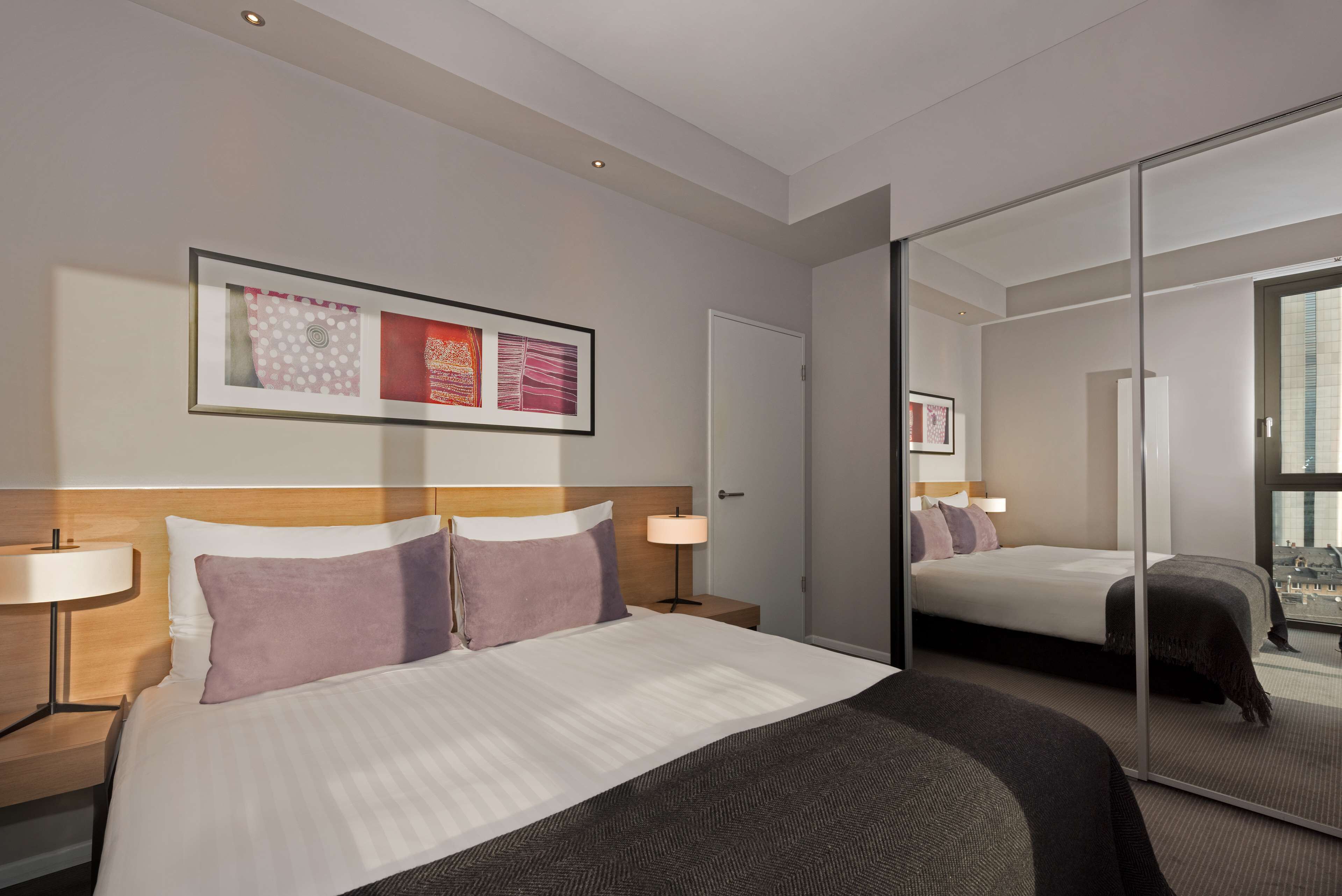 Habitación de hotel con una cama grande, una mesita de noche con lámpara, una obra de arte enmarcada sobre la cama y un ventanal corredero que da a un balcón.
