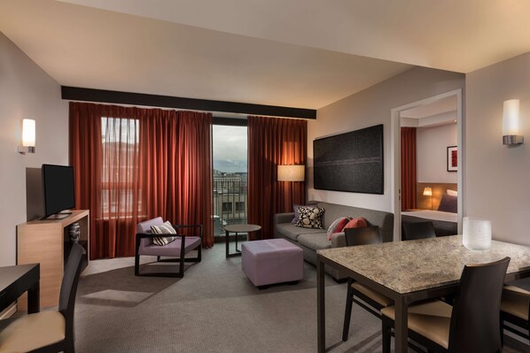 Habitación de hotel con zona de estar, televisión, mesa de comedor y vistas a la ciudad a través de amplios ventanales.