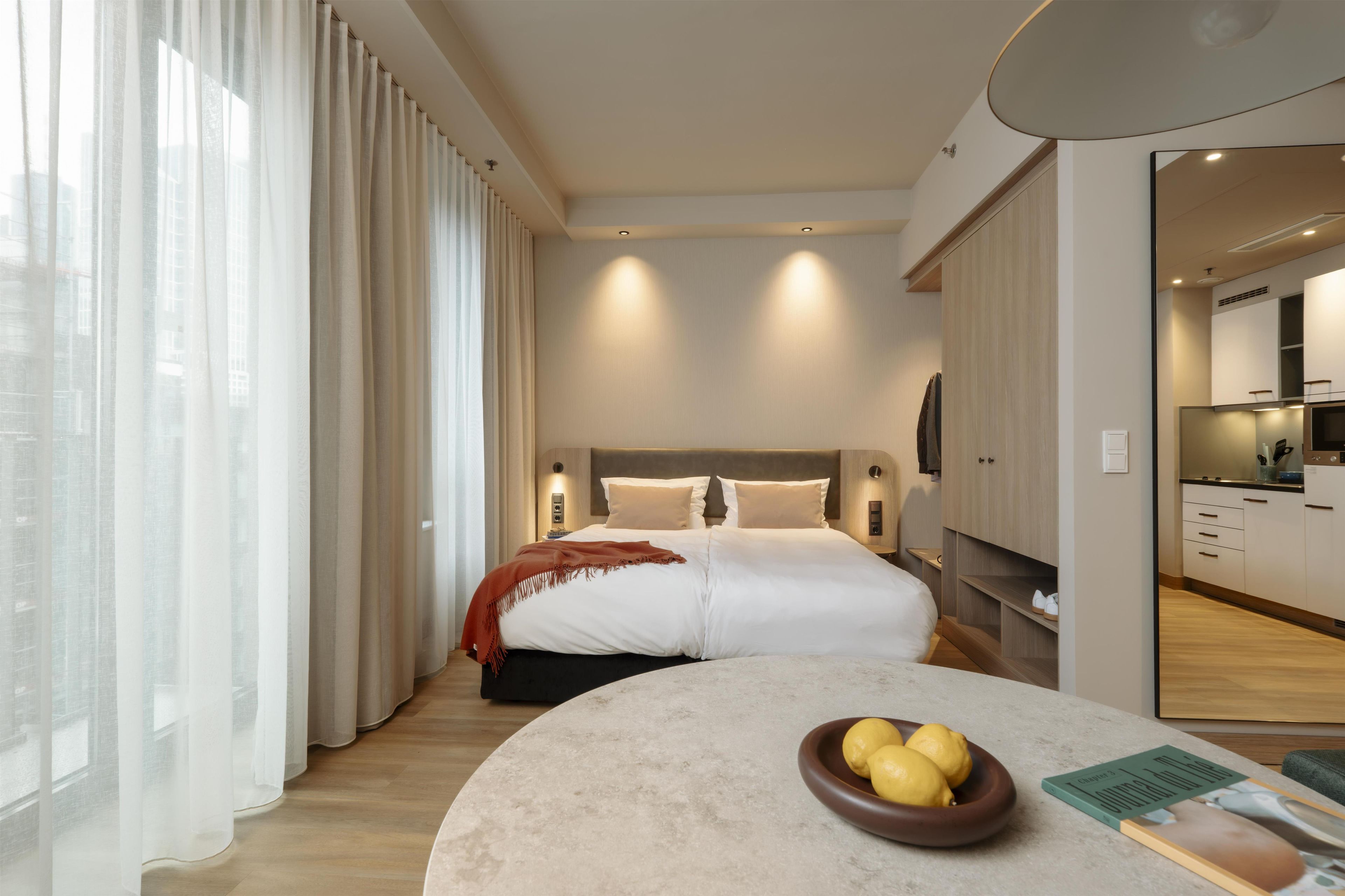 Ein modernes Hotelzimmer mit einem großen Bett, einem runden Tisch mit einer Zitronenschale und Blick auf die Stadt durch transparente Vorhänge.