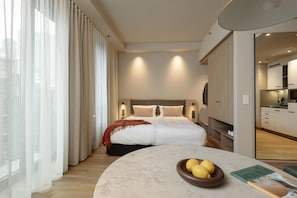 Ein modernes Hotelzimmer mit einem großen Bett, einem runden Tisch mit einer Zitronenschale und Blick auf die Stadt durch transparente Vorhänge.