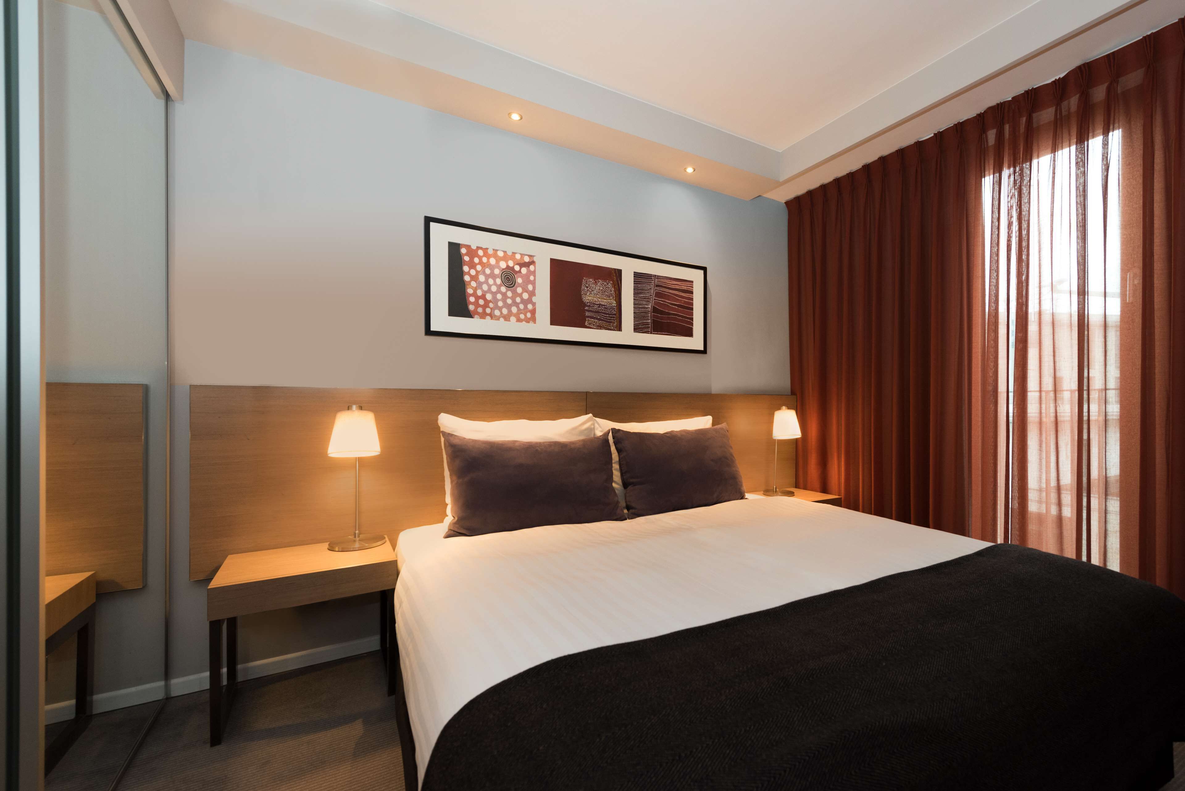 Een hotelkamer met een groot bed, twee bedlampen, een kunstwerk in een lijst boven het bed en een raam met gordijnen.