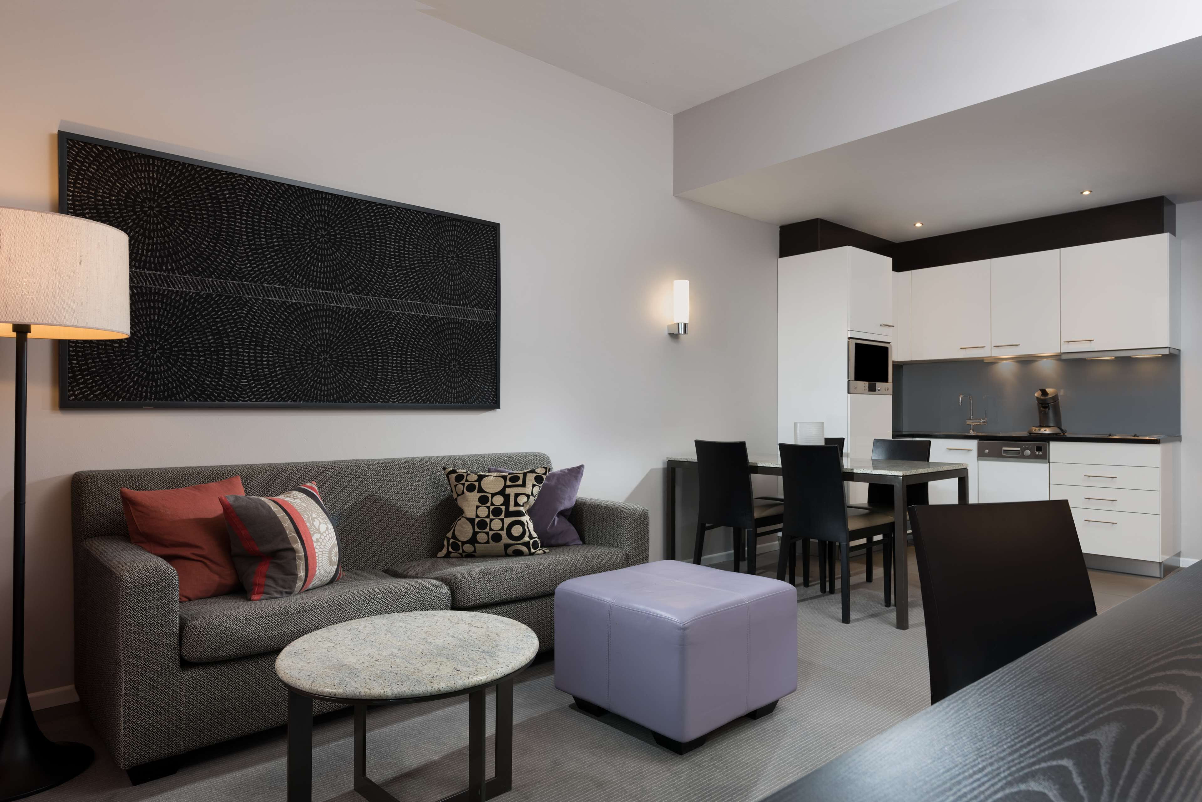 A modern living room with a grey sofa, a round coffee table, and a purple ottoman. There is a dining area with a table and chairs, and a kitchen with white cabinets and a black countertop.