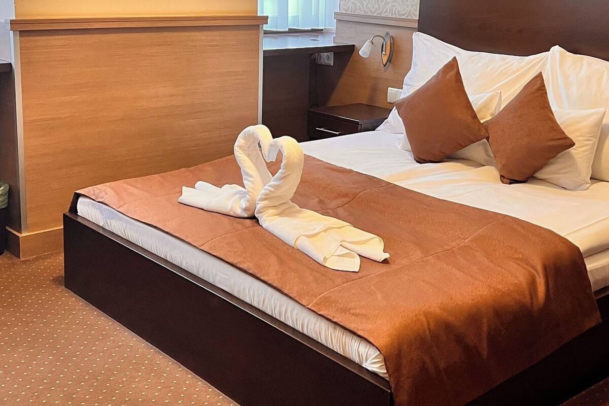 A hotel room with a large bed, a wooden headboard, a nightstand, a wall clock, and a window with curtains.