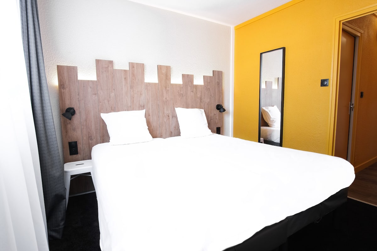 Une chambre d’hôtel avec une tête de lit en bois, un lit avec du linge de lit blanc, une petite table de chevet, un miroir et une porte sur le côté droit.