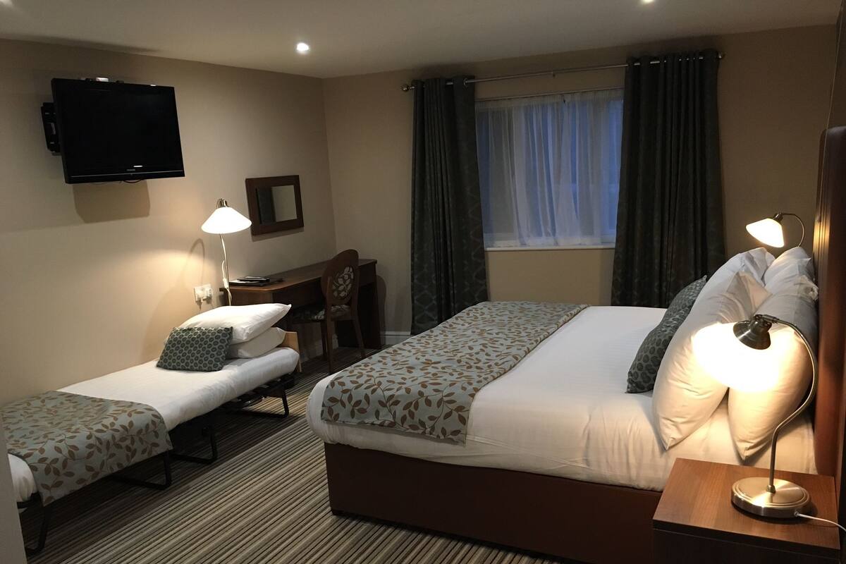 A hotel room with two single beds, a larger bed, a desk with a chair, a TV mounted on the wall, and a window with curtains.