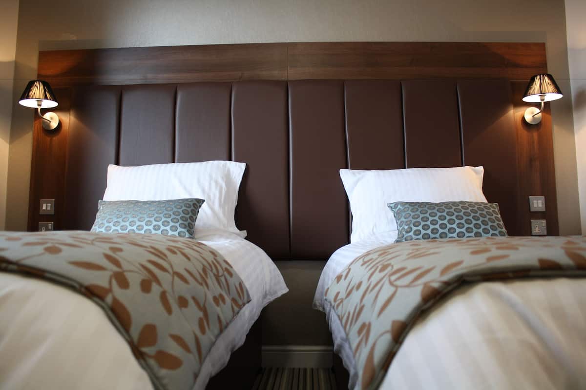 A hotel room with two beds, each with a patterned bedspread and matching pillows. There are wall-mounted lamps on either side of the beds.