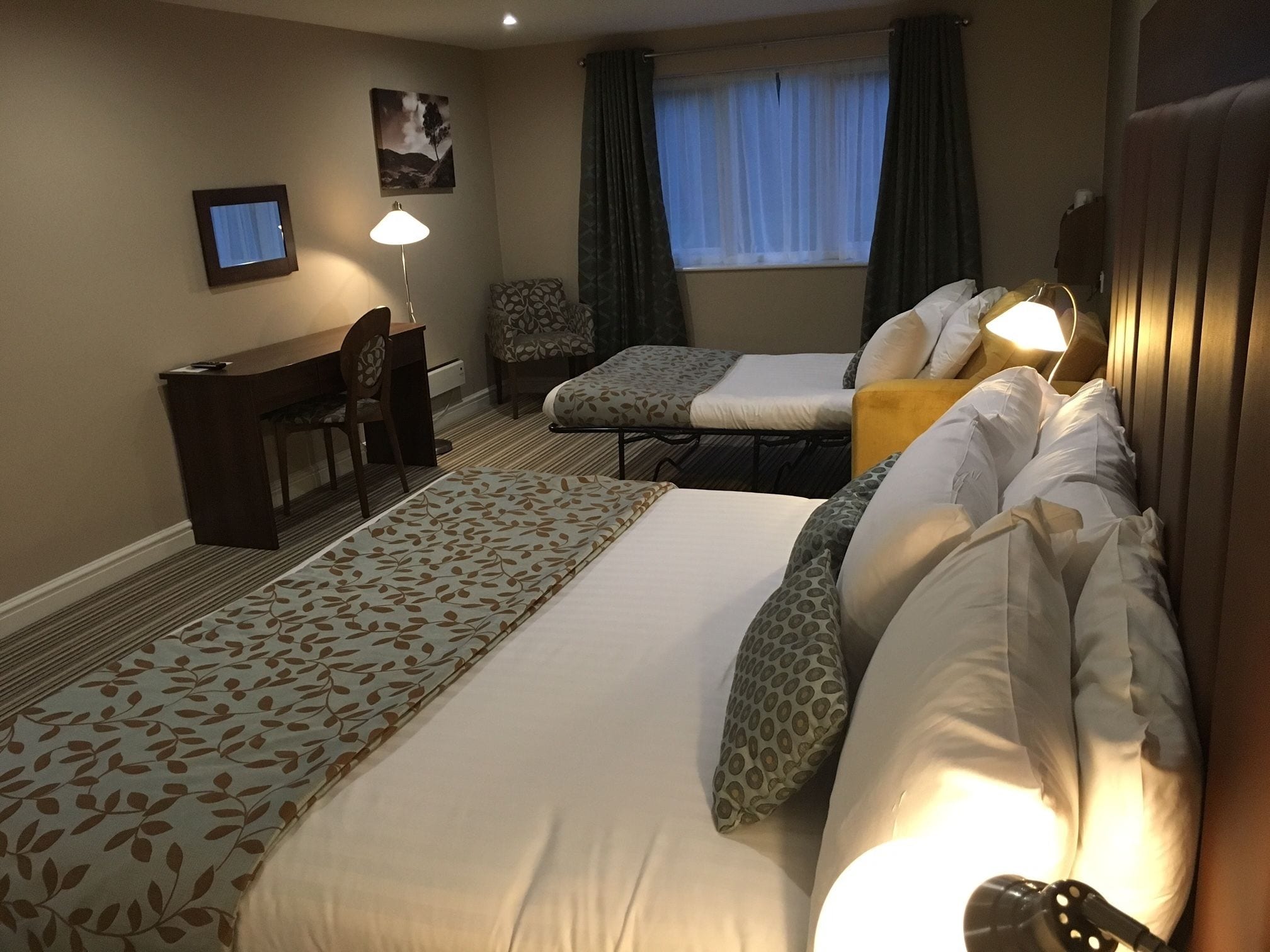 A hotel room with a large bed, a smaller bed, a desk, a chair, a lamp, a window with curtains, and a painting on the wall.
