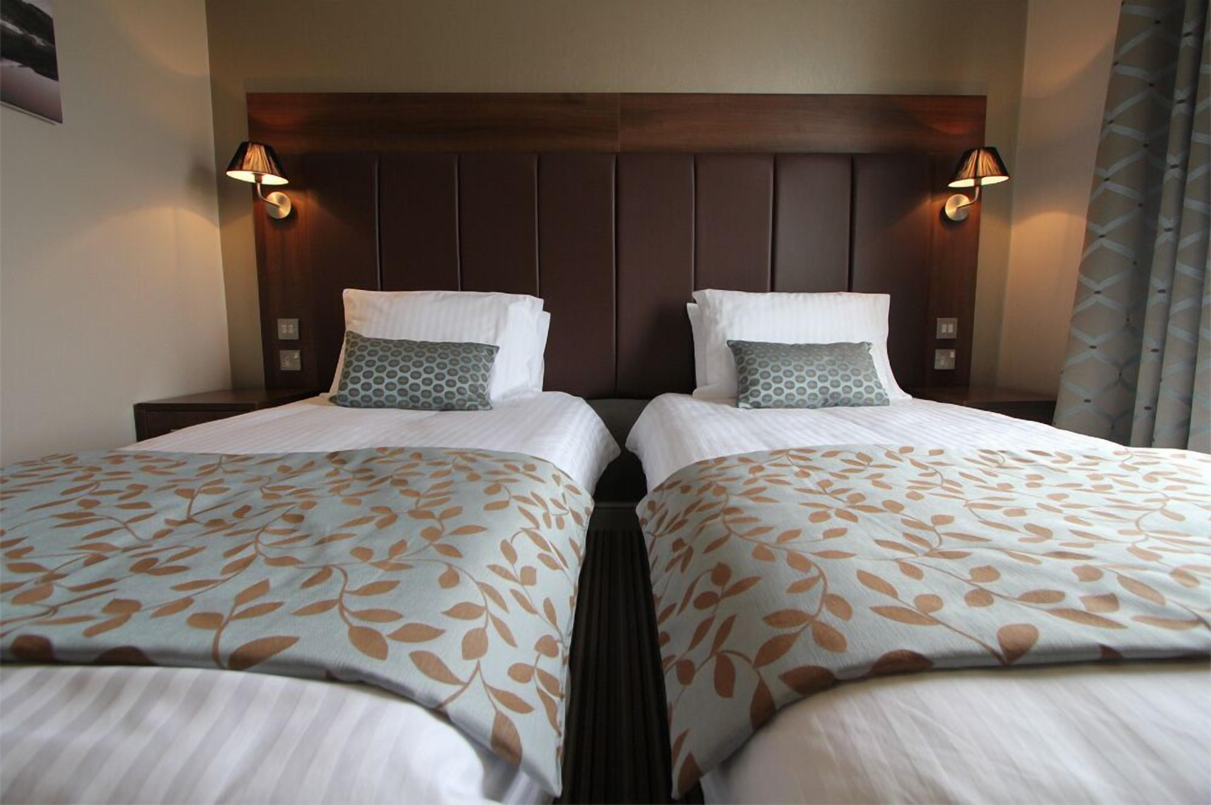 A hotel room with two beds, each with a patterned bedspread and pillows, a wooden headboard, and wall-mounted lamps.