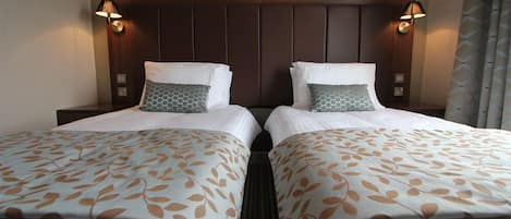 Habitación de hotel con dos camas, cada una con una colcha y cojines estampados, cabeceros de madera y lámparas fijadas a la pared.