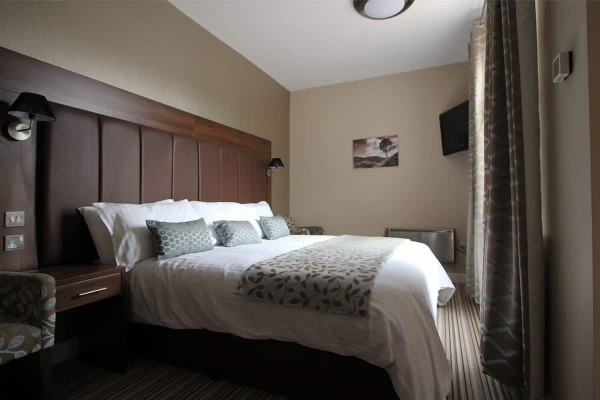 A hotel room with a large bed, a headboard, a bedside table, a lamp, a painting on the wall, and a window with curtains.