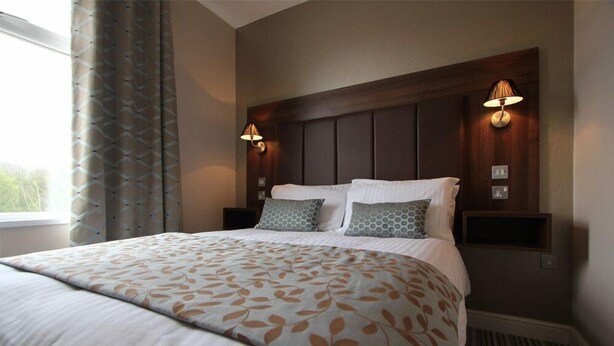 A hotel room with a large bed, two bedside lamps, a headboard, and a window with curtains. - Twin Oaks Hotel (Chesterfield)