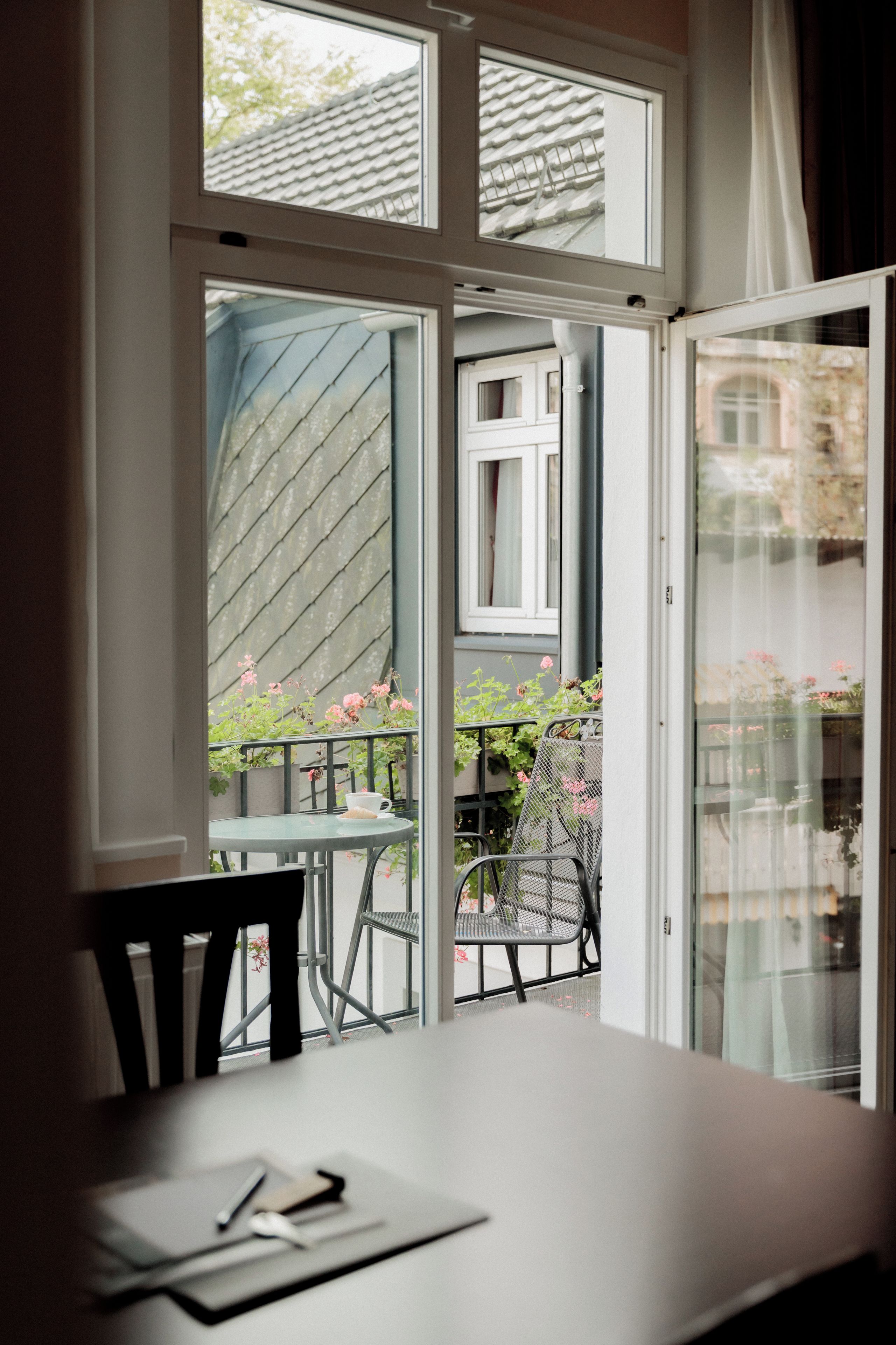 Eine offene Tür führt zu einem Balkon mit Tisch und Stühlen, ein Fenster mit Blick auf ein Gebäude und ein Stuhl im Inneren.