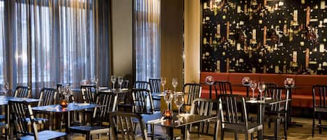 A restaurant with tables set for dining, featuring a modern interior with a patterned wall and large windows with curtains.