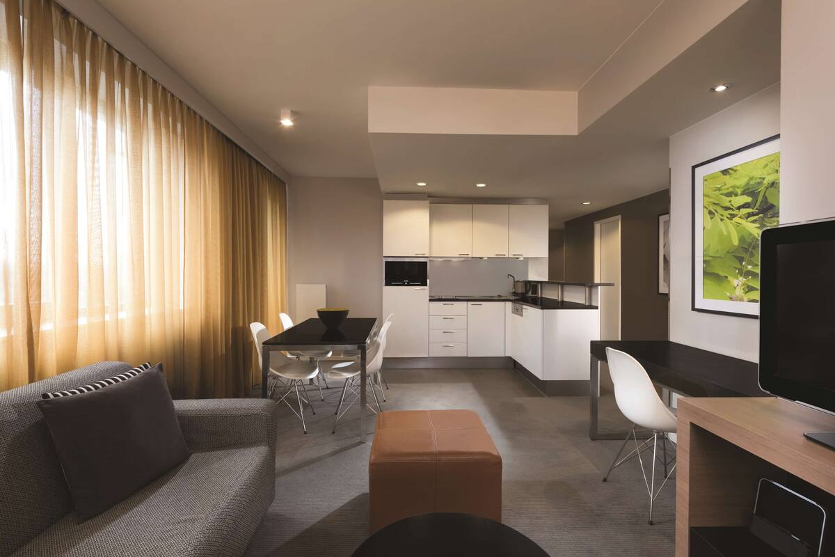 Ein modernes Hotelzimmer mit Sofa, einem Esstisch mit Stühlen, einer Küchenzeile und einem Fernseher.