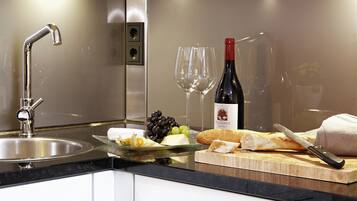 A kitchen counter with a stainless steel sink, a bottle of wine, a glass, a cutting board with cheese, grapes, and a baguette.