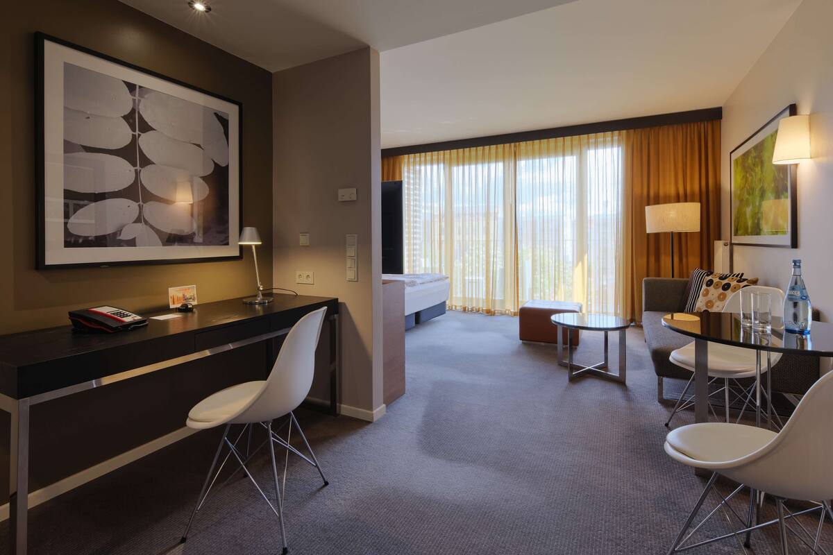 Ein modernes Hotelzimmer mit Schreibtisch, Stühlen, einem Bett und Blick auf die Stadt durch große Fenster.