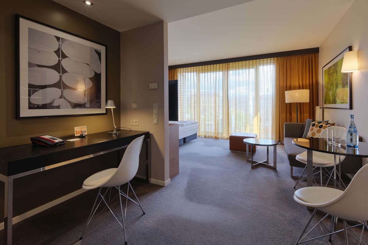 Une chambre d’hôtel moderne équipée d’un bureau, de chaises, d’un lit et offrant une vue sur la ville grâce à de grandes fenêtres.