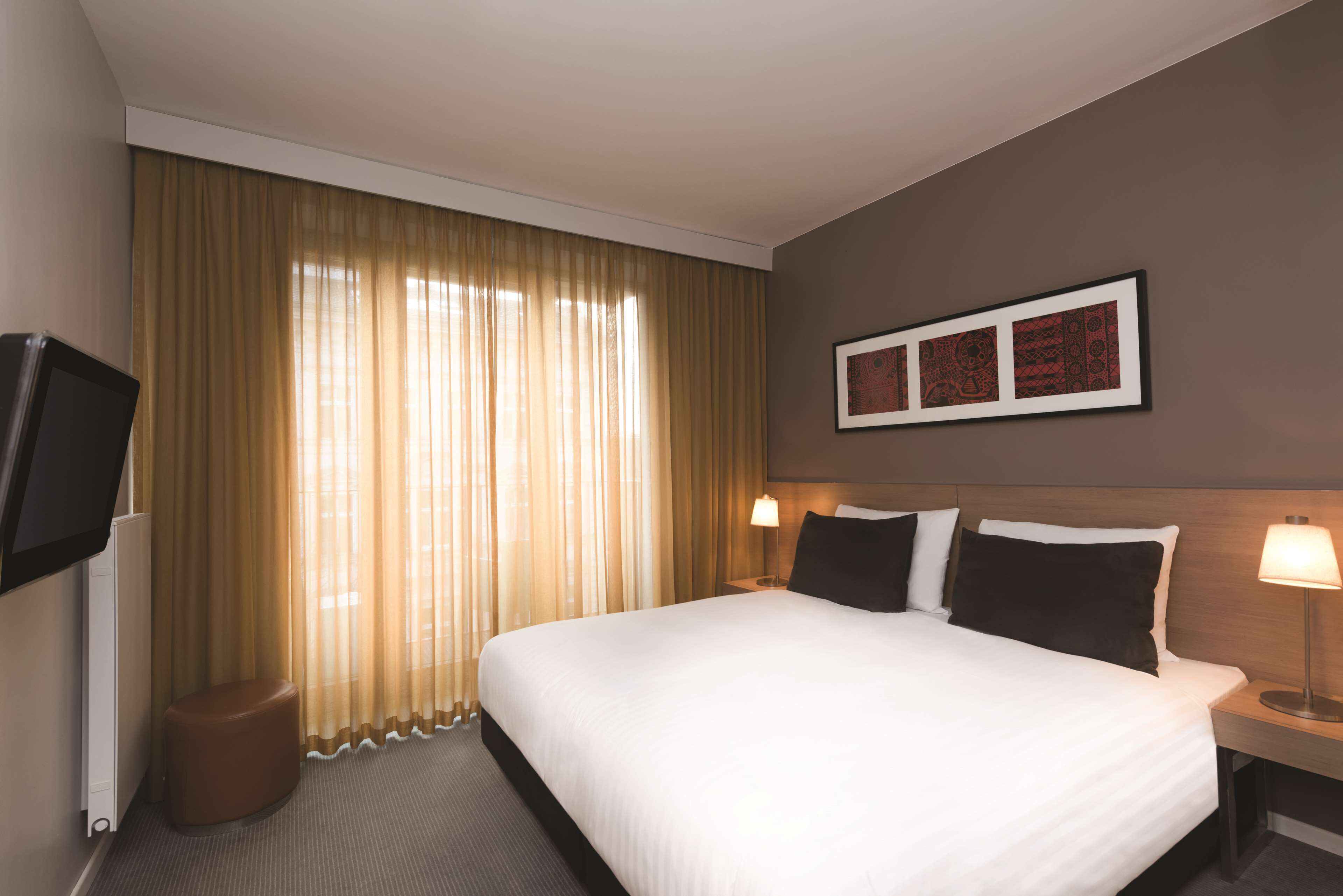 Une chambre d’hôtel avec un grand lit, un téléviseur à écran plat fixé au mur, une table de chevet avec une lampe et un tableau encadré au-dessus du lit.