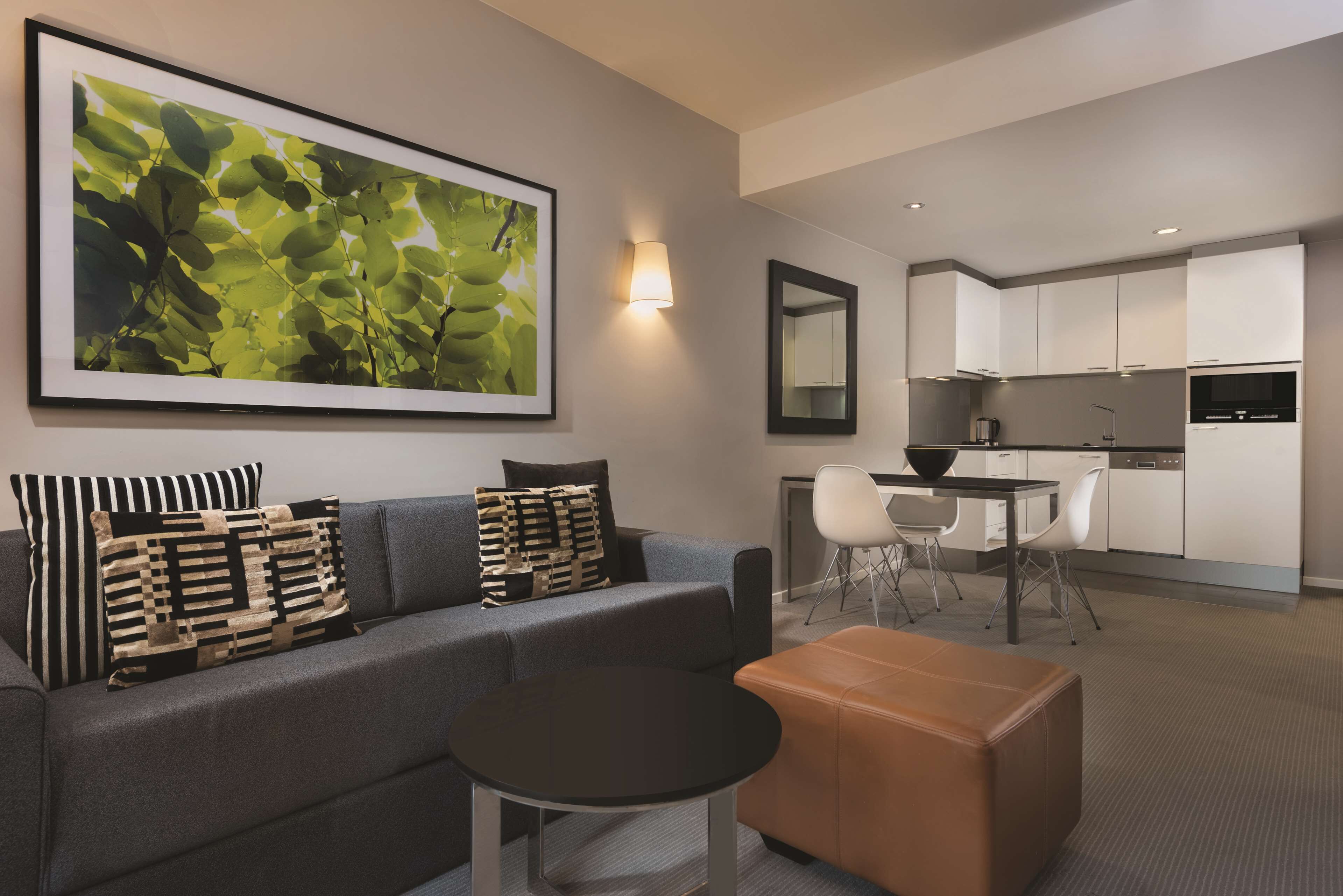 Un salon moderne avec un canapé gris, une table basse noire et un pouf marron. Un grand tableau encadré orne le mur, et on y trouve également un coin cuisine avec des meubles blancs, ainsi qu’une table à manger avec des chaises.