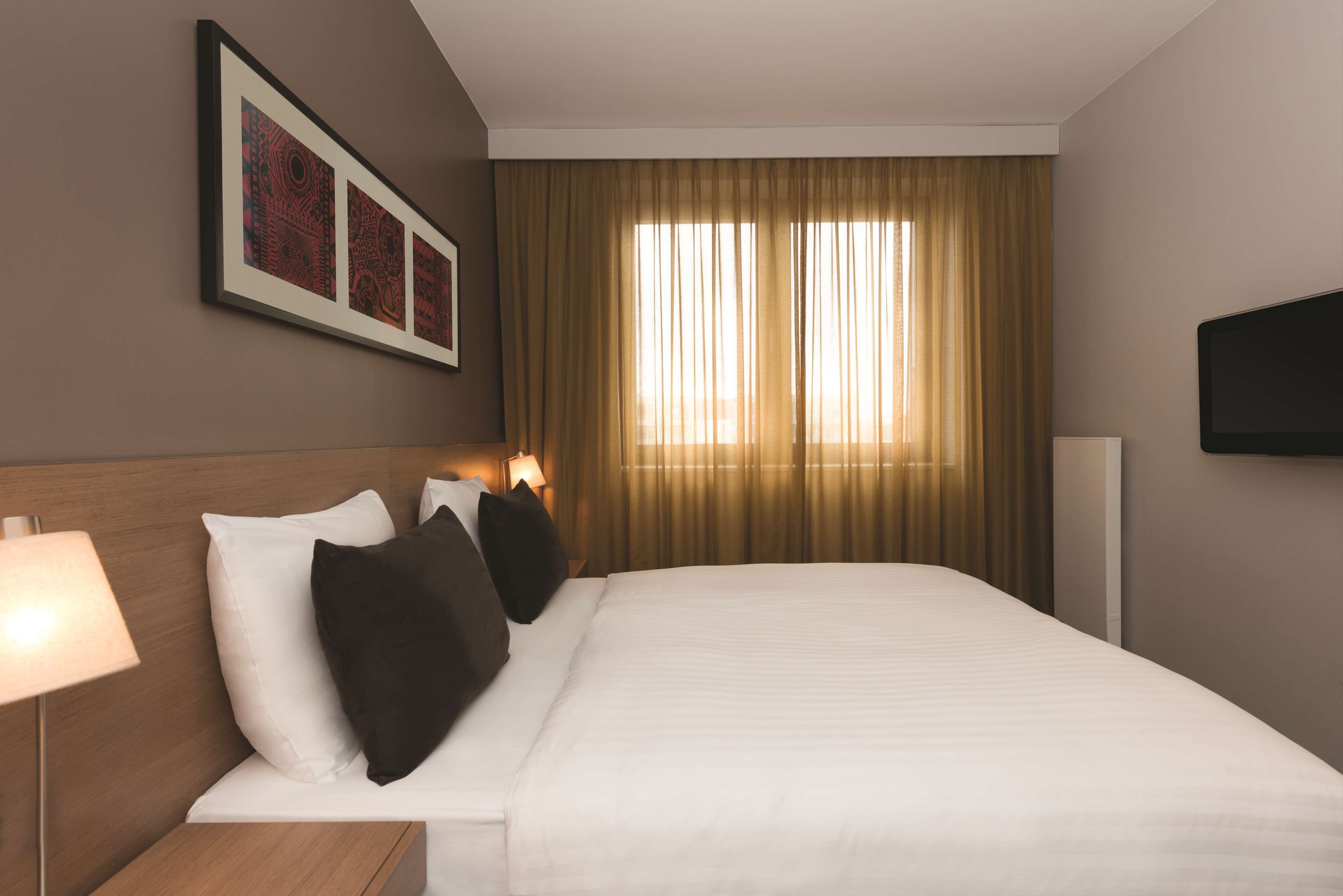 A hotel room with a large bed, a flat-screen TV mounted on the wall, and a window with curtains.