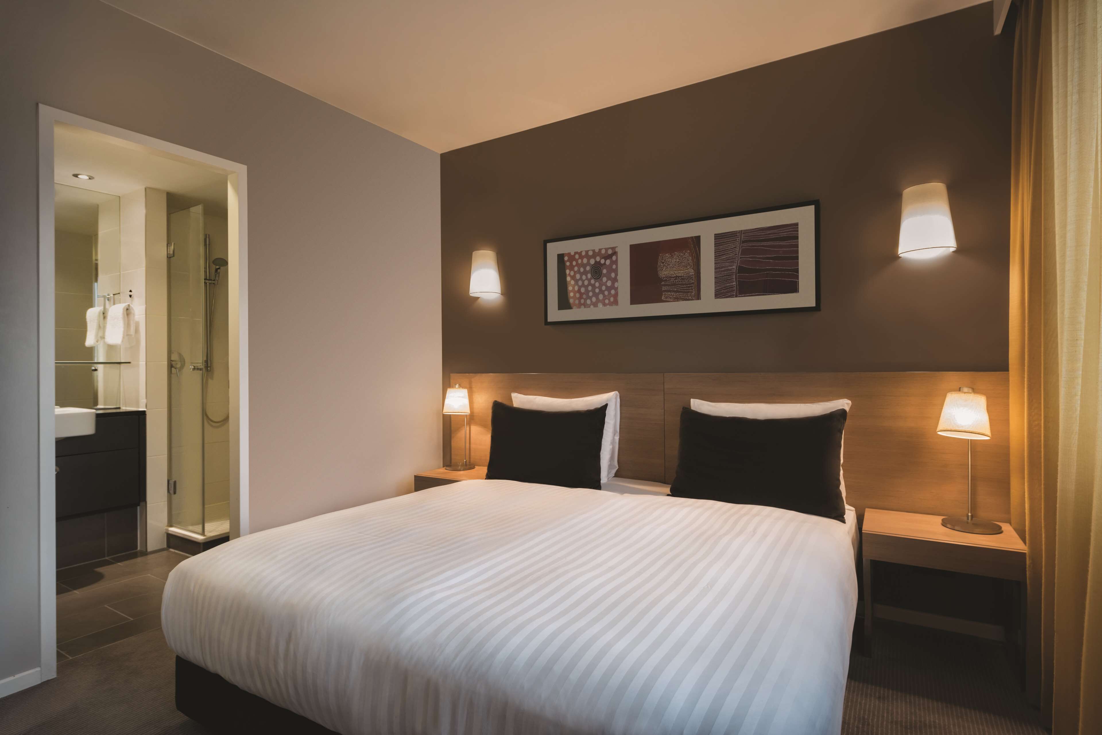 A hotel room with a large bed, two bedside tables with lamps, a framed picture on the wall, and a bathroom visible through an open door.