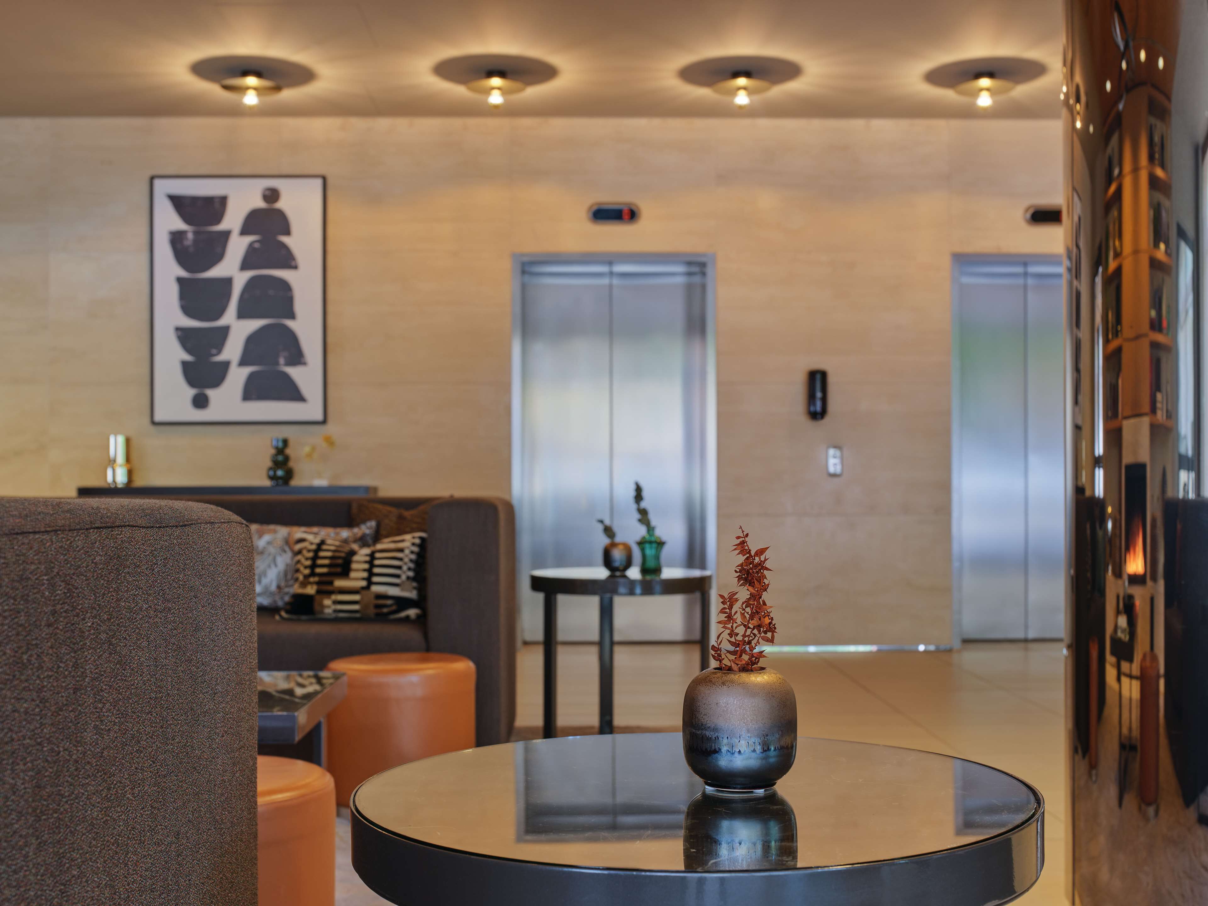 Un hall d’entrée moderne avec un canapé, un pouf et une table ronde agrémentée d’un vase. On aperçoit des ascenseurs au fond.
